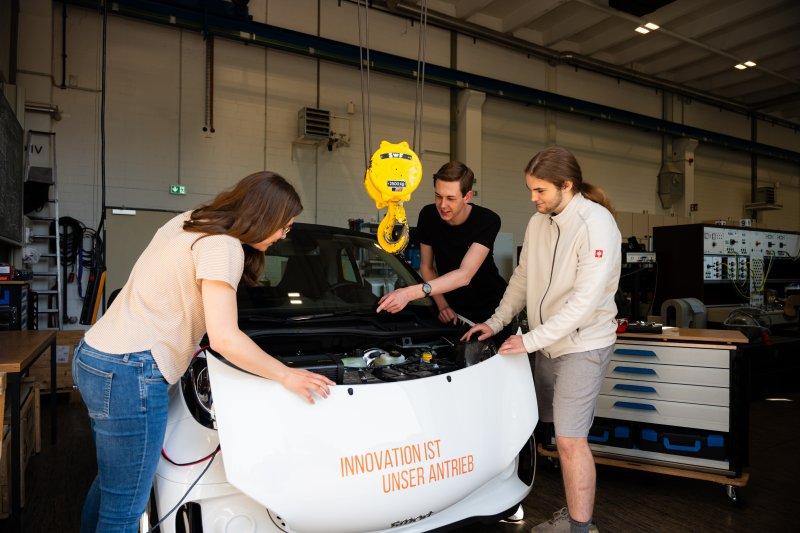 Zwei junge Männer und eine junge Frau stehen an der geöffneten Motorhaube eines E-Smarts, auf der Motorhaube steht: Innovation ist unser Antrieb.