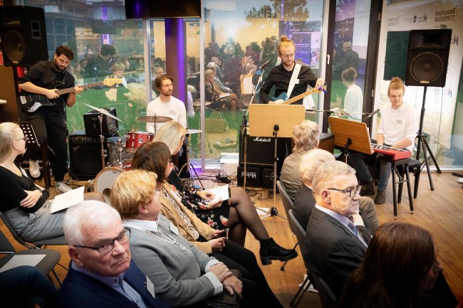 Publikum und dahinter eine Band, hinter den Musikern spiegelt die Glasscheibe den Raum.