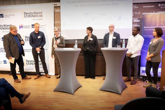 Eine Gruppe Menschen hinter Stehtischen, einige halten Mikrofone. Im Bildrand ist Publikum zu erkennen.