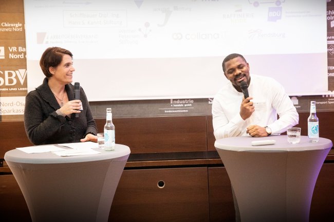 Eine Frau und ein Mann an Stehtischen im Gespräch, beide halten ein Mikro und lachen.