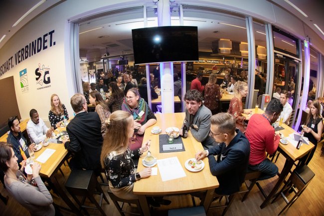 Menschen sitzen an einem Tisch und unterhalten sich, in der Fensterscheibe im Hintergrund spiegelt sich der Raum.