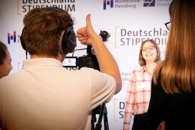 Ein junger Mann hinter einer Kamera, eine junge Frau davor. Er hebt den Daumen.