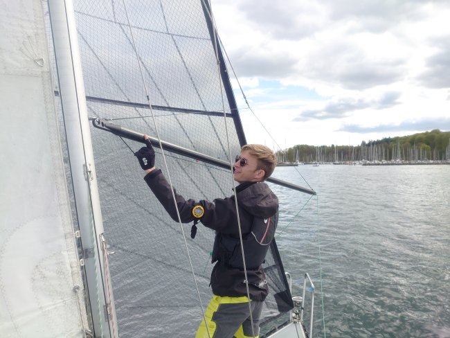 Ein junger Mann in sportlicher Kleidung steht auf einem Segelboot und verändert etwas an der Einstellung des Segels.