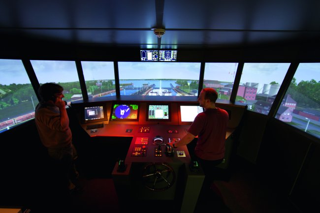 Ein dunkler Raum, in dem die Gerätschaften einer Schiffsbrücke aufgebaut sind - dahinter sieht man die Projektion einer Schleuse. Zwei junge Männer hantieren mit den Gerätschaften.