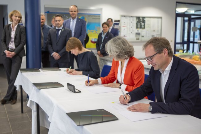Ein Mann und zwei Frauen setzen ihre Unterschrift unter drei Dokumente, im Hintergrund sieht man eine Gruppe von Menschen in Anzügen, die sich die Unterzeichnung anschauen.