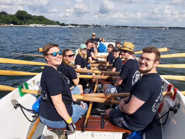 Eine Gruppe von jungen Menschen sitzt in einem breiten Kutter mit eingelegten Riemen und lächelt in die Kamera. Im Hintergrund sieht man Wasser und das Ufer.