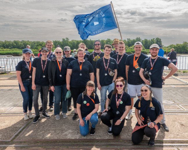 Eine Gruppe von jungen Menschen steht auf einer Hafenpromenade und lächelt in die Kamera. Einer schwenkt eine blaue Flagge mit der Aufschrift &quot;Kutterpullen Hochschule Flensburg&quot;.