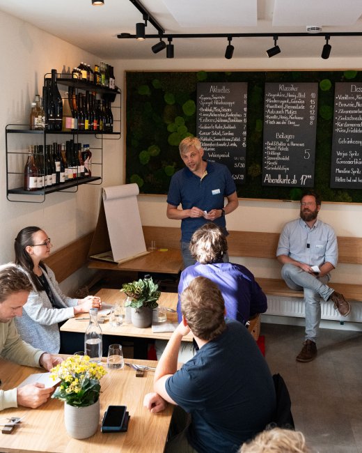 Kneipe: Im Hintergrund eine Tafel mit Angebiten, vorn besetzte Tische, vor der Tafel steht ein mann und spricht.