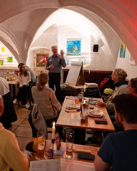 Kneipe: Besetzte Tische in einem gewölbeartigen Keller, vorn steht ein Mann und spricht.