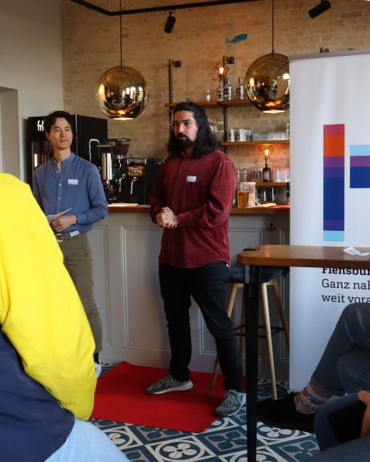 Zwei Männer stehen mit dem Rücken zu einer Bar und erzählen etwas.