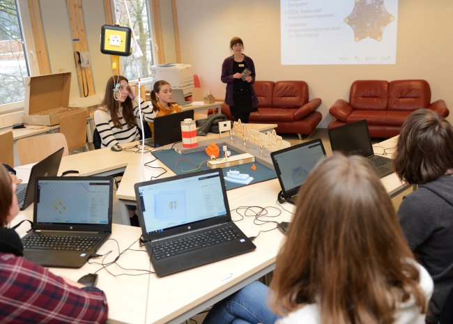 Blick über die Schulter eines Mädchens, vor ihr ein Laptop. Davor im Raum ein Modell eines Hafens und eine weibliche Lehrkraft.