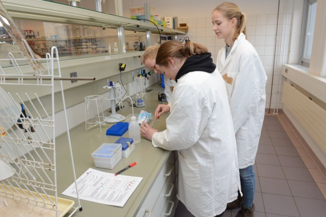 Drei Schülerinnen in Laborkitteln im Labor. Man sieht sie eher von hinten.