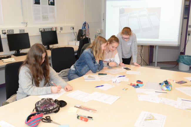 Vier Schülerinnen arbeiten an einem Tisch. Vor ihnen liegen Papier, Stifte und Handys.