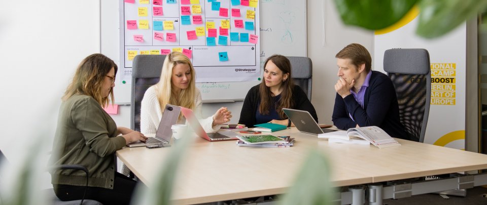 Eine Gruppe von Menschen sitzt um einen Arbeitstisch, hinter ihnen an der Wand hängt ein Whiteboard voller bunter Post Its, vor ihnen auf dem Tisch Laptops, Bücher und Stifte.