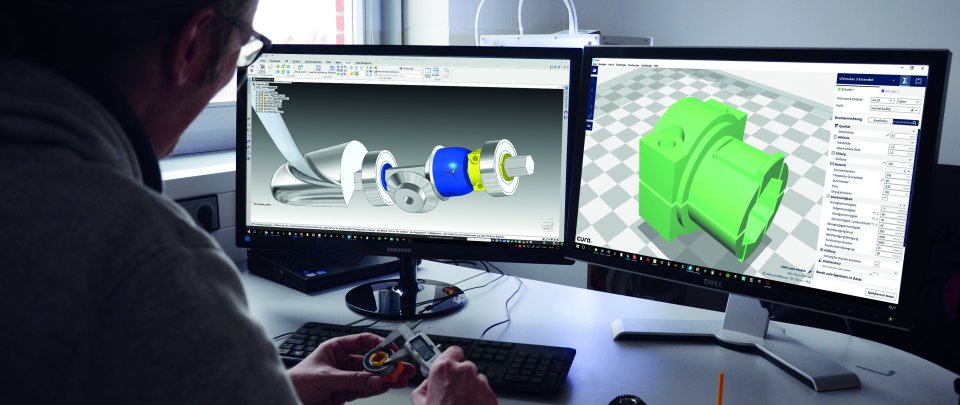 Mensch sitzt vor zwei Computerbildschirmen. Fokus des Bildes sind die Bildschirme, auf denen 3D-Modellierungen zu erkennen sind.