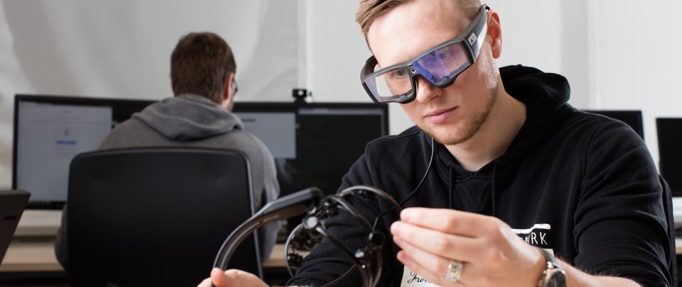 Student mit Brille und Headset an einem Schreibtisch. Im Hintergrund ist ein anderer student an einem Computer sitzend zu erkennen.