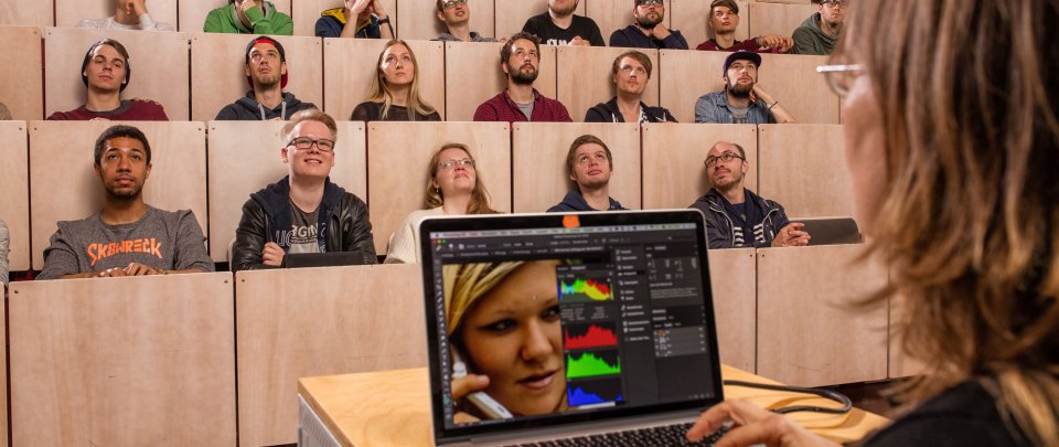 Studierende im Hörsaal. Vorn, eher rechts im Bild und angeschnitten der Hinterkopf einer Dozentin, die einen Laptop bedient.