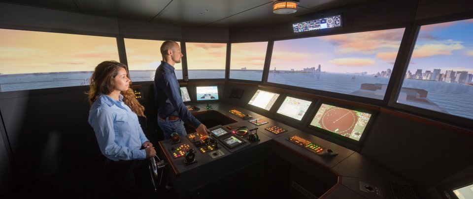 Studierende im Schiffsführungssimulator. Vor ihnen beleuchetete Bedienungspanel, durch die Fenster der Schiffbrücke ist eine Szene auf See zu erkennen.