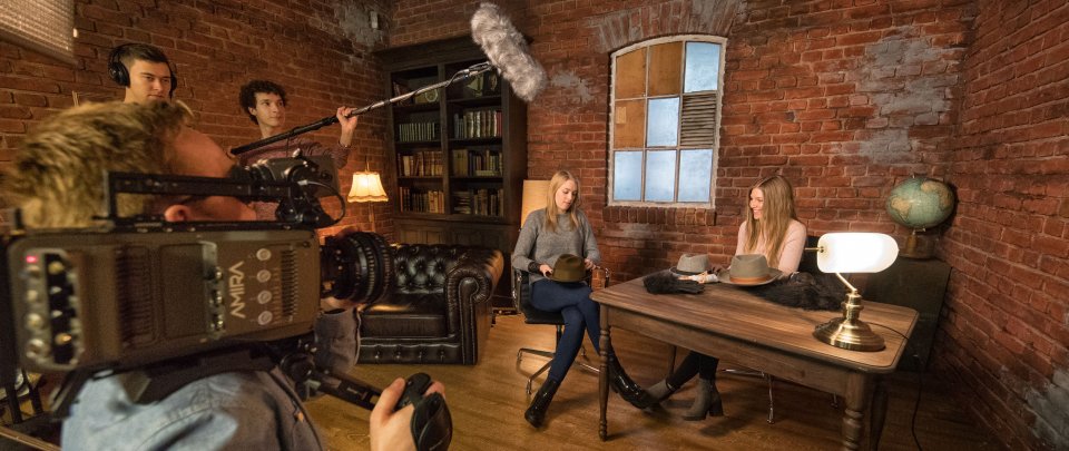 Students filming in the university's TV studio. You can see a camera and sound equipment and two female students sitting on stage.