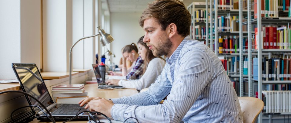 Student arbeitet in der Bibliothek am Laptop. Im Hintergrund andere Studierende und Bücherregale.