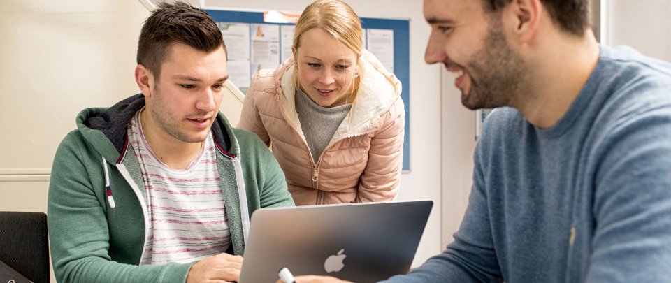 3 Studierende in einer Lernsituation, zwei blicken gemeinsam auf einen Laptop.