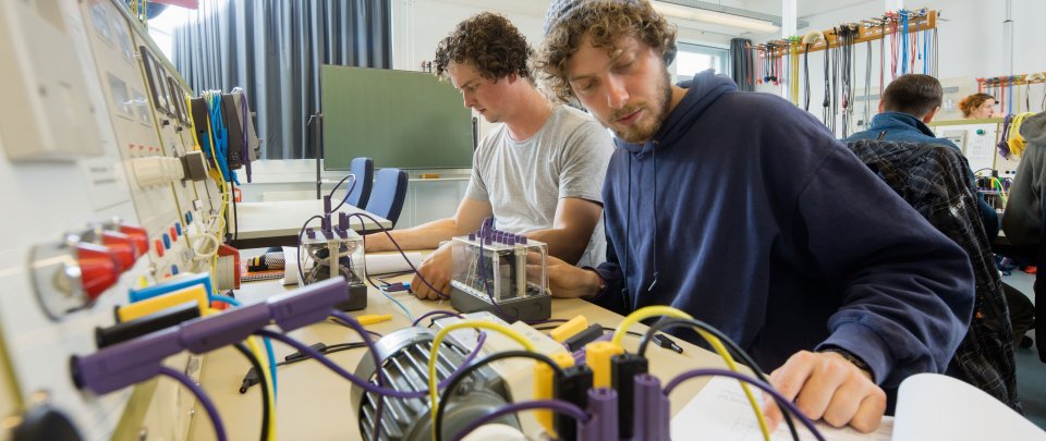 Zwei Studenten beim Arbeiten im Labor, im Bildvordergrund diverse Kabel und Arbeitsunterlagen, hinten sind andere Studierende zu erkennen.