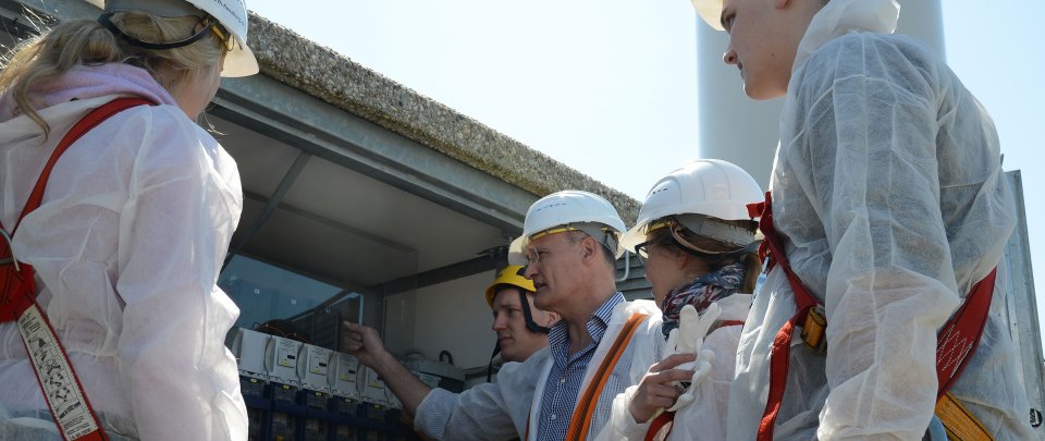 Professor und Studierende in Arbeitsschutzkleidung am Fuß einer Windenergieanlage. Der Professor zeigt die darin verborgene Technik.