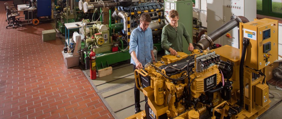 Zwei Studenten in der Maschinenhalle arbeiten an einer Baumaschine. Im Hintergrund sind weitere große Maschinen zu erkennen.