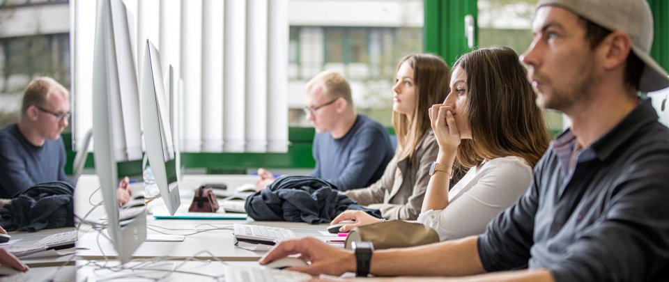 Rechts im Bild eine Reihe Studierender, die nebeneinander sitzen und je auf einen Computerbildschirm schauen. Zum Teil werden sie links im Bild in Bildschiormen gespiegelt.