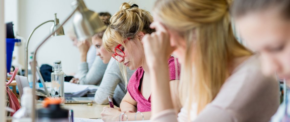 Mehrere Studierende, die in einer Reihe an einem langen Tisch sitzen und über Bücher gebeugt sind oder am Laptop arbeiten. Einige sind unscharf zu erkennen, den Bildmittelpunkt bildet eine blonde Frau mit auffällig roter Brille.