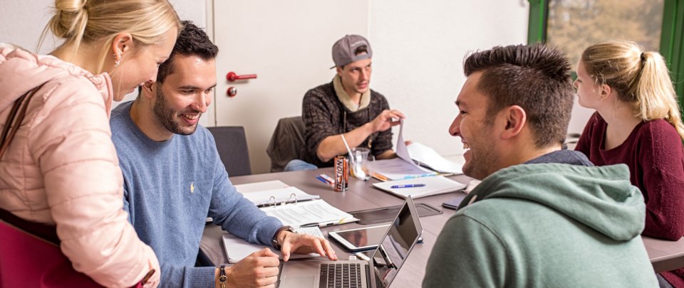 Eine Gruppe Studierender sitzt um einen Tisch. Einige sind im Gespräch miteinander, andere schauen auf einen Laptop.