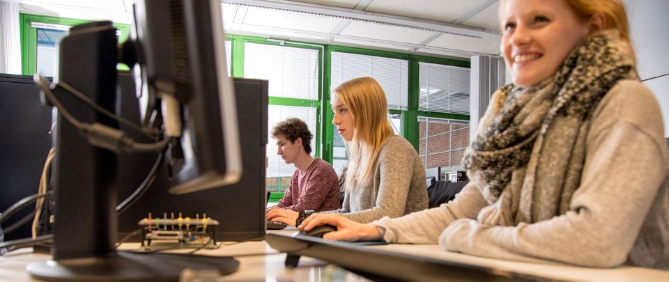 Zwei Studentinnen und ein Student sitzen nebeneinander je an einem Rechner in einem Computerlabor. Die Studentin vorn im Bild läöchelt, vor ihr auf dem Tisch liegt außer Maus und Tastatur ein Konstrukt mit diversen Kabeln uns´d Steckern.