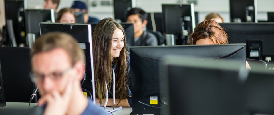 Blick auf ein Computerlabor, in dem mehrere Studierende an Rechnern arbeiten. In der ersten Reihe ist unscharf ein blonder Student zu erkennen, im Fokus in der Reihe hinter ihm eine dunkelhaarige, lachende Studentin.