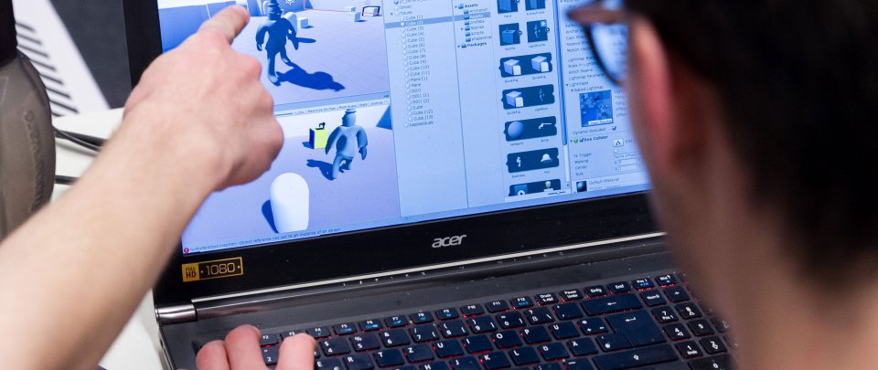 Wir blicken einem kurzhaarigen Menschen mit Brille über die Schulter, der vor einem Bildschirm sitzt, auf dem das Entwicklungsstadium ein Computerspiel zu erkennen ist. Seine Hände liegen auf der Tastatur unten im Bild. Von links ragt ein Arm ins Bild, der auf den Bildschirm zeigt.