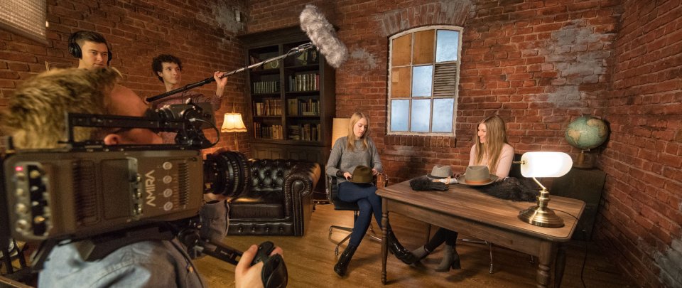 Eine Gruppe Studierender im TV-Studio: Vorn links hält ein Student eine Kamera, neben ihm halten zwei weitere Studenten ein Mikrofon, einer trägt Kopfhörer. An einem Tisch rechts im Bild sitzen zwei Studentinnen in einer Interviewsituation. Vor ihnen auf dem Tisch liegen Hüte und andere Requisiten. Der Raum ist mit Ledersofa, Bücherregal und Globus eingerichtet.