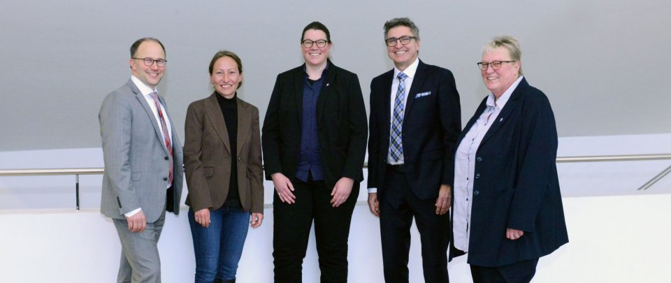 Das Präsidium vor einer schrägen weißen Wand im audimax. Die Gruppe besteht aus fünf Menschen, alle tragen eher formelle Kleidung und lächeln in die Kamera.