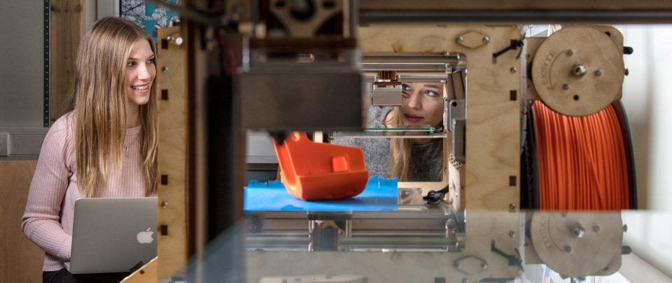 Zwei Frauen arbeiten an einem §D-Drucker, eine hat einen Laptop auf dem Schoß. Der 3D-Drucker ist im Bildvordergrund.