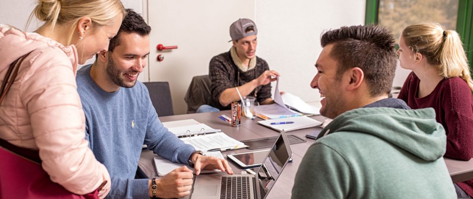 Gruppe Studierender um einen Tisch versammelt, lernend und im Gespräch.