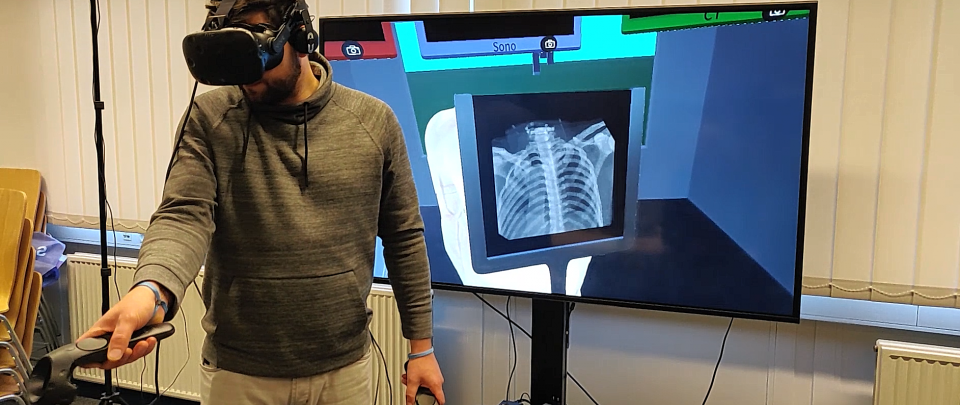 Person mit Virtual Reality-Brille im Vordergrund. Die Person hält einen und Virtual Reality-Controller in der Hand. Im Hintegrund ist ein großer Monitor zu sehen, auf dem eine virtuelle 3D-Szene dargestellt wird, in der ein Röntgenbild zu sehen ist.