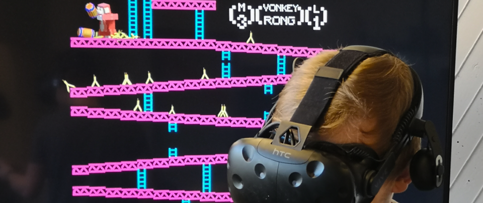 Im Vordergrund ist der Kopf einer Person zu sehen, die eine Virtual Reality-Brille trägt. Im Hintergrund sieht man auf einem großen Computermonitor eine Szene aus einem Computerspiel, das Donkey Kong ähnelt.