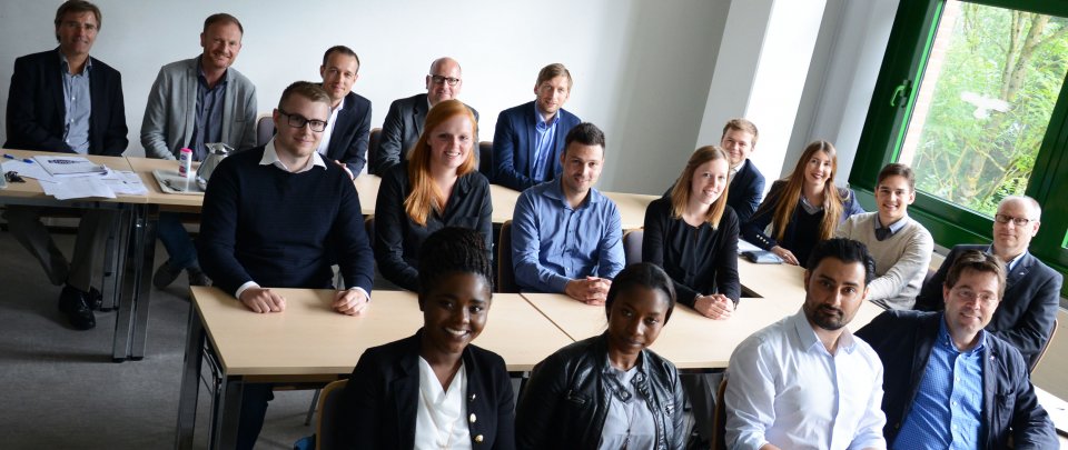 Gruzppe von Studierenden und Lehrenden sitzen an Tischen in einem Seminarraum und gucken in die Kamera.