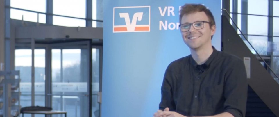 Junger Mann in schwarzem Oberteil steht vor einem VR Bank-Banner und schaut lächelnd in die Kamera.