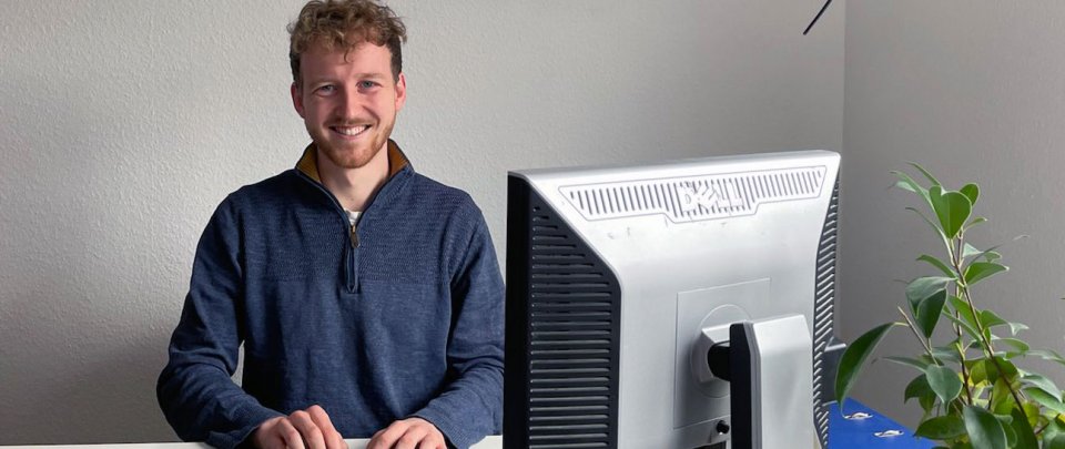 Junger Mann sitzt am Schreibtisch und lächelt in die Kamera. Vor ihm ein Computerbildschirm und eine Zimmerpflanze.