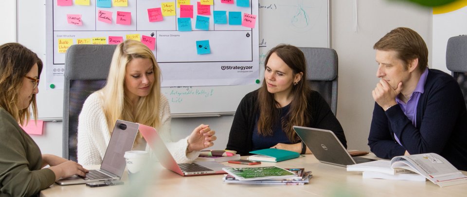 Eine Gruppe von Menschen sitzt um einen Arbeitstisch, hinter ihnen an der Wand hängt ein Whiteboard voller bunter Post Its, vor ihnen auf dem Tisch Laptops, Bücher und Stifte.