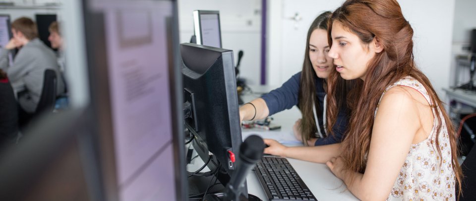 Zwei junge Frauen von der Seite fotografiert: Sie sitzen vor einem Computer, vorn im Bild ein Mikrofon.