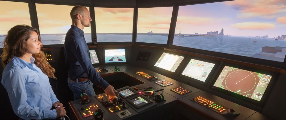 Studierende im Schiffsführungssimulator. Vor ihnen beleuchetete Bedienungspanel, durch die Fenster der Schiffbrücke ist eine Szene auf See zu erkennen.
