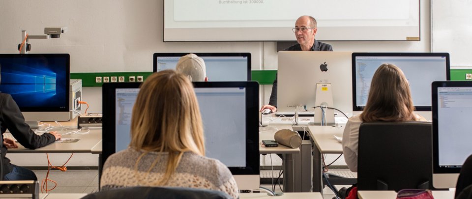 Blick von einen Studierenden auf seine Mitstudierenden, die vor Ihren PC sitzen und einem Dozenten zuhören
