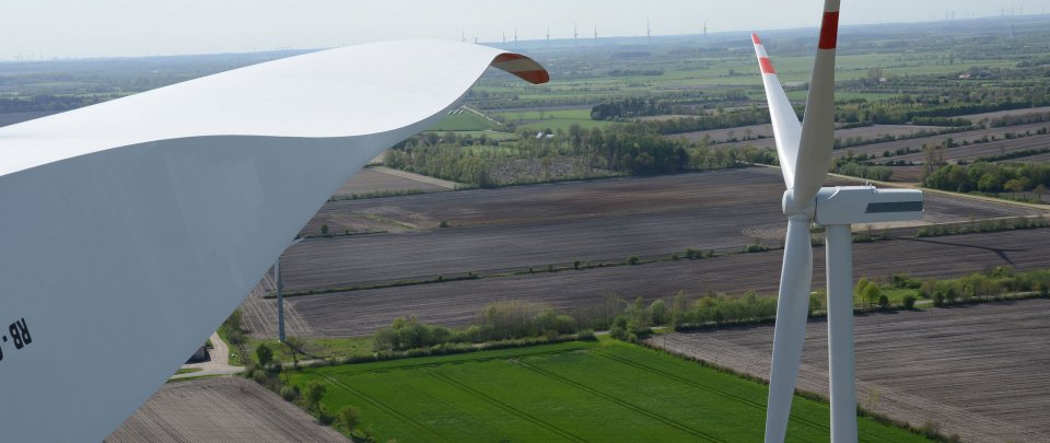 Windkraftanlage, im Hintergrund Felder und Himmel, vorn im Bild unscharf Rotorblatt einer anderen Anlage.