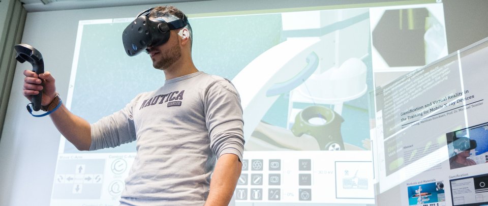 Mann steht vor einer Wand, auf der ein virtuelles Röntgengerät zu erkennen ist. Er trägt eine VR-Brille und hält einen Controller vor sich.