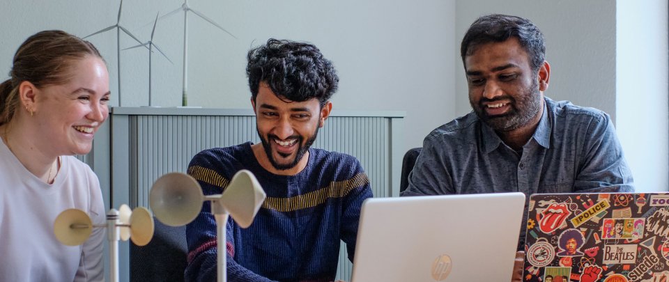 Drei Studierende arbeiten an Laptops, hinter und vor ihnen Modelle von Windkraftanlagen. Sie lachen.